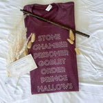 Load image into Gallery viewer, Flat lay image of Heather Maroon Tee Shirt that reads: Stone, Chamber, Prisoner, Goblet, Order, Prince, Hallows in white block lettering. Styled with wand and Fall themed plants. 
