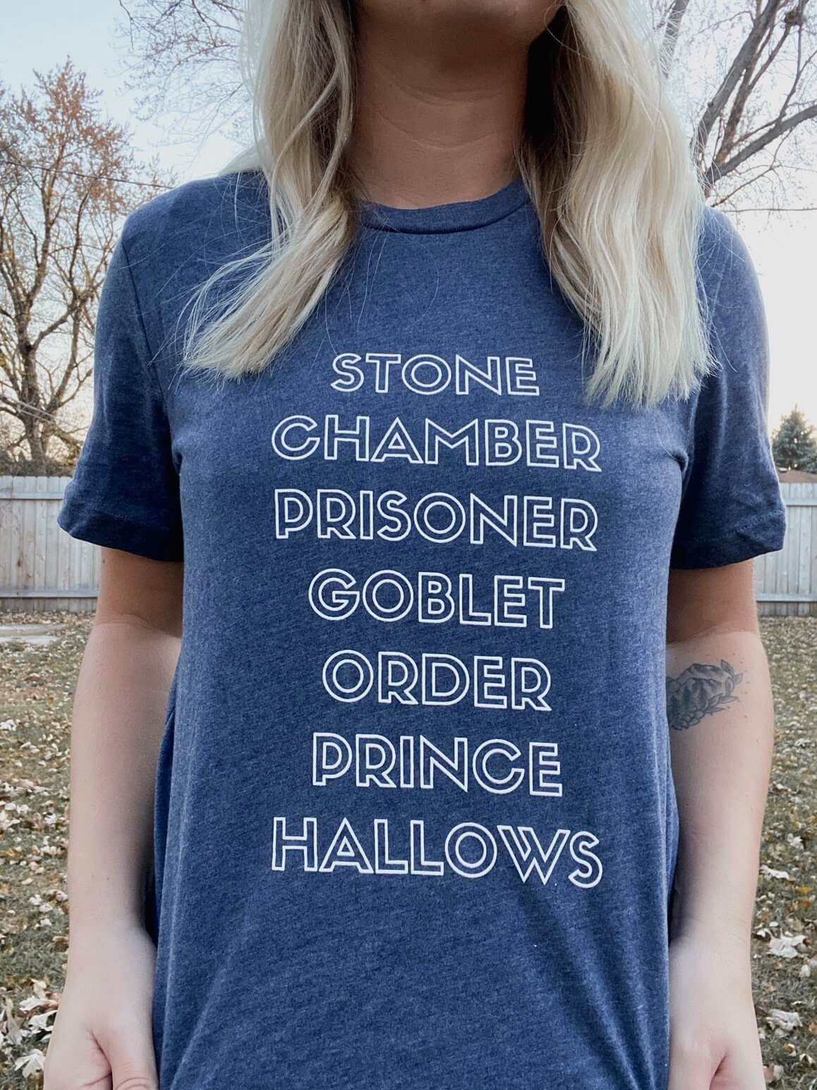 Woman wearing Heather Navy Tee Shirt that reads: Stone, Chamber, Prisoner, Goblet, Order, Prince, Hallows in white block lettering.