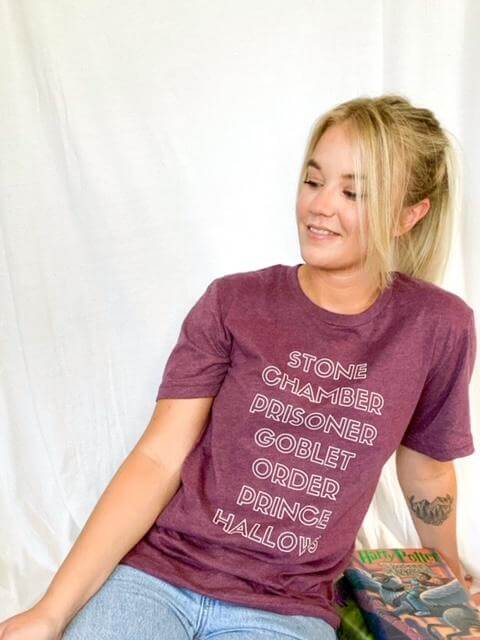 Woman wearing Heather Maroon Tee Shirt that reads: Stone, Chamber, Prisoner, Goblet, Order, Prince, Hallows in white block lettering.