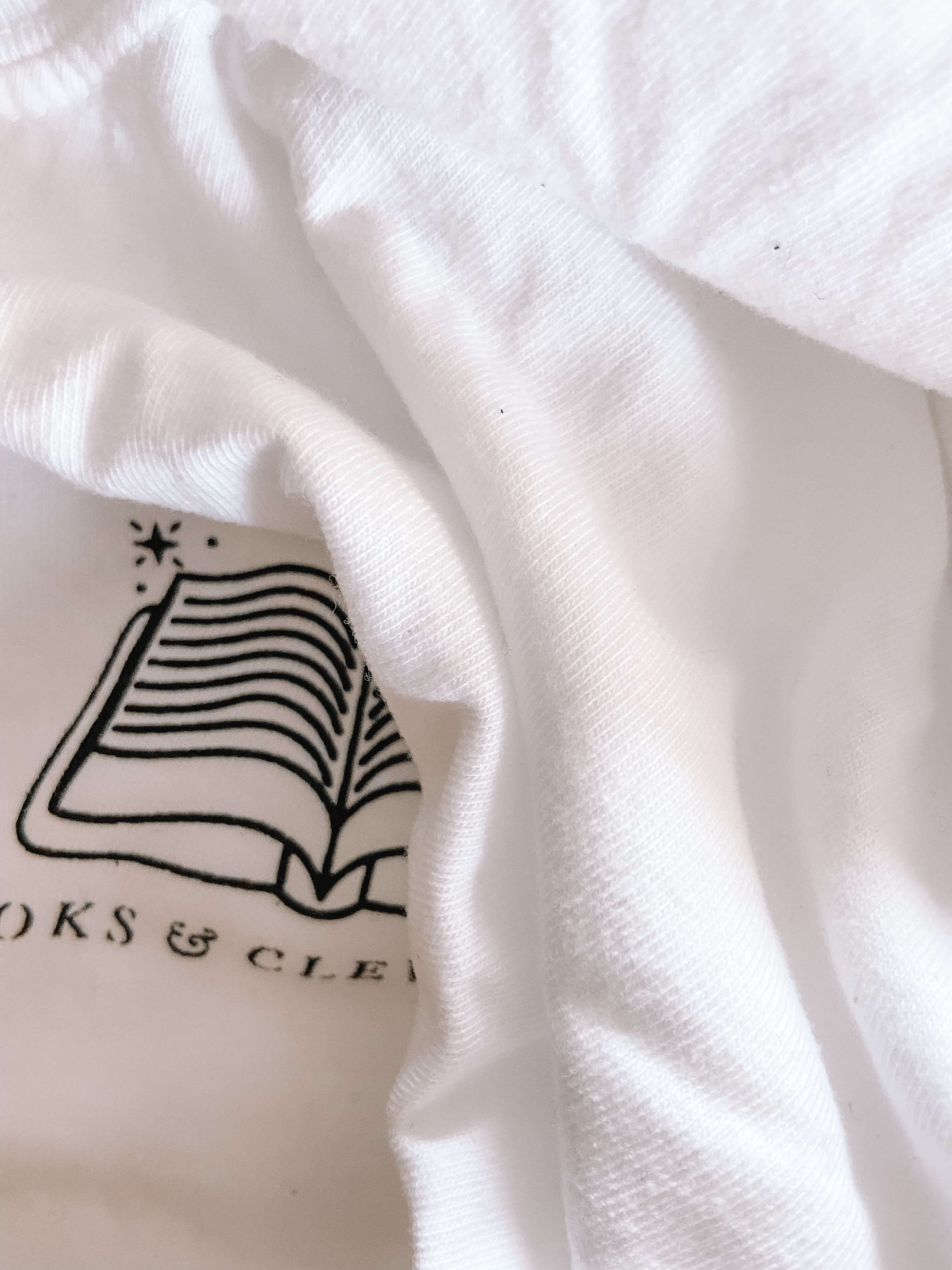 Closeup of white tee shirt with black logo that reads "Books and Cleverness" with art of an open book above it. 