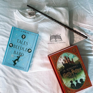 Flat Lay of white tee shirt with black logo on the left chest. Logo reads "Books and Cleverness" with clip art of an open book above it. Styled with a wand and 1 Tales of Beedle the Bard book and one Harry Potter and the Sorcerer's Stone book. 