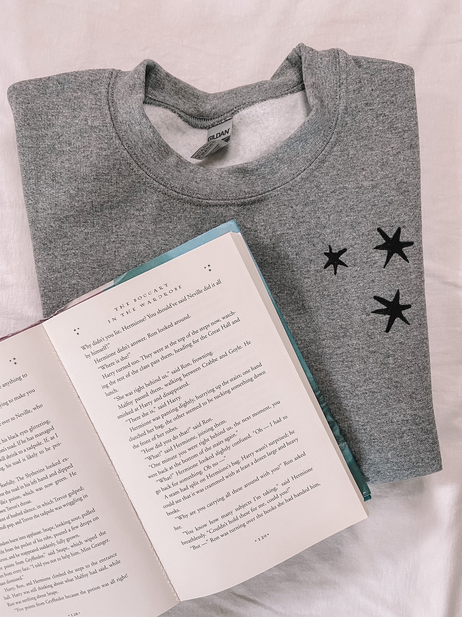 Flat lay of charcoal gray crew neck sweatshirt that has 3 black embroidered stars on the left chest. Styled with an open Harry Potter and the Prisoner of Azkaban book.